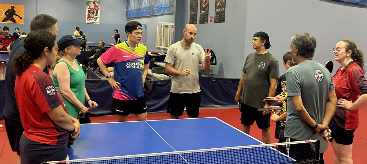A coach leading a group lesson with players gathered around the table at Princeton Pong