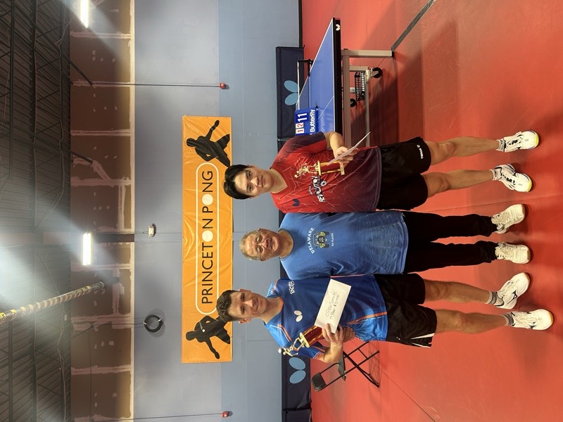 Two players holding trophies and a first-place prize envelope with a club coach, standing under the Princeton Pong banner